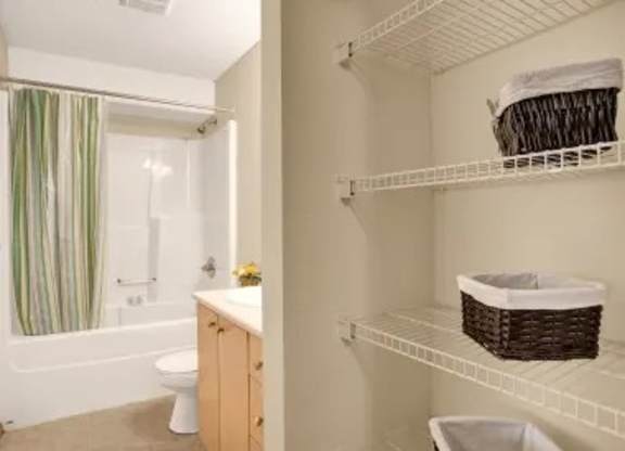 A clean bathroom here at Wellington Manor with a bathtub-shower combo, a simple striped shower curtain, and a white toilet. A light wood vanity with a built-in sink adds a warm and practical touch. A spacious open shelving area with wire racks provides convenient storage for towels and everyday essentials.