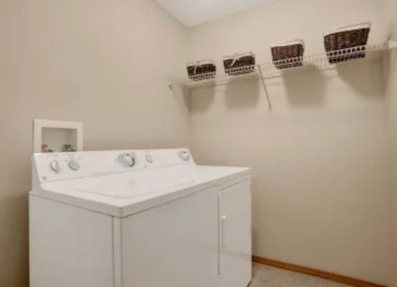 A convenient laundry area here at Wellington Manor with a full-size washer and dryer and simple neutral walls. A long wire shelf above provides extra space for laundry baskets and supplies. A clean, functional setup makes everyday routines easy and organized.