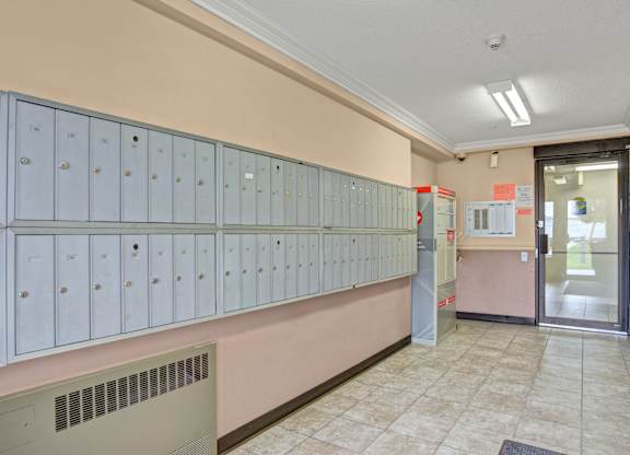 A clean community mailroom here at Wellington Manor with a long wall of secure resident mailboxes and bright overhead lighting. A tiled floor and neutral wall tones create a neat and well-kept space. A convenient entry door and parcel area provide easy access for everyday mail pickup.
