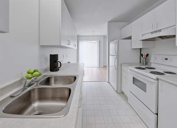 A clean galley-style kitchen here at Willowview Terrace with white cabinets, a double stainless-steel sink, and bright countertops for a simple modern look. A white refrigerator and electric stove sit along the right side, with light flooring and a dining area visible near the sliding glass doors at the back.