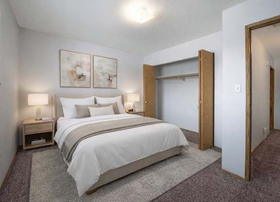 A bright, cozy bedroom here at Willowview Terrace featuring a neatly made bed with white bedding and soft neutral accent pillows. Matching nightstands with simple lamps sit on each side, while two modern wall art pieces add a clean touch above the headboard. An open closet and plush carpet complete the simple, comfortable layout.