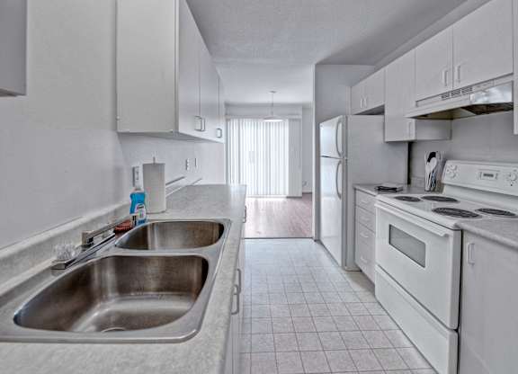 A bright galley-style kitchen here at Willowview Terrace with white cabinets, light countertops, and a double stainless steel sink. White appliances including a stove, range hood, and refrigerator line the space, with durable tile flooring that leads into the dining area with a sliding glass door for natural light.