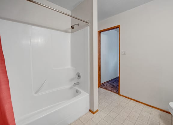 A bright bathroom tub and shower area here at Willowview Terrace with smooth white surround walls, a simple showerhead, and a sturdy curtain rod. Light tile flooring and warm wood trim lead out to the hallway, keeping the space clean, open, and easy to maintain.