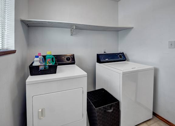 A convenient laundry area here at Willowview Terrace with a full-size washer and dryer, a wide storage shelf for supplies, and a nearby window that brings in natural light. A simple basket setup adds extra organization for everyday laundry needs.