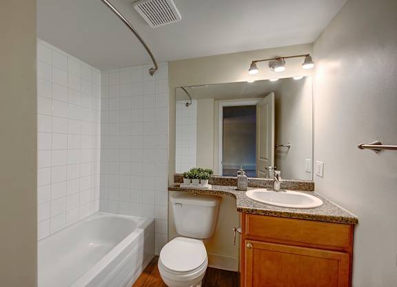 A clean bathroom here at Wyndham Crossing with a white tub and shower surround, a bright vanity light, and a wide wall mirror for an open feel. A wood-tone sink cabinet with a speckled countertop and simple towel bars add a practical touch to the space.