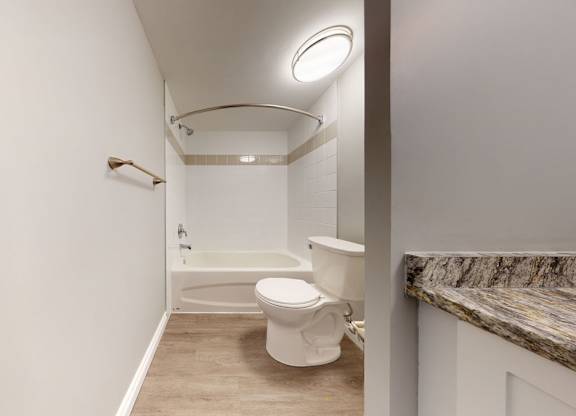 A simple bathroom here at Wyndham Crossing featuring a white tub and shower combination, a curved shower rod, and bright overhead lighting. A white toilet sits beside the tub, while wood-look flooring and a granite-style countertop add a clean, modern finish.