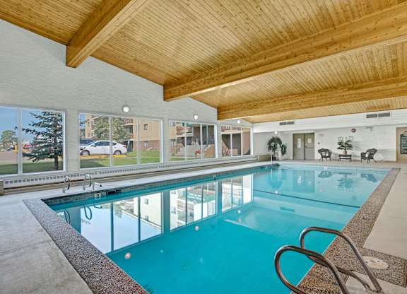 A bright indoor swimming pool here at Wyndham Crossing with clear blue water, a wide deck area, and stainless steel handrails for easy access. Large windows line the wall to bring in natural light, and the warm wood-beam ceiling adds a welcoming touch to the space.