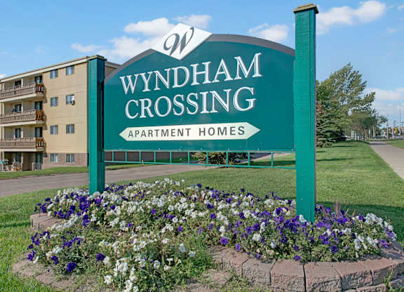 A welcoming community entrance sign here at Wyndham Crossing featuring a large green “Wyndham Crossing Apartment Homes” display surrounded by a colorful flower bed. A multi-story apartment building and neatly kept green lawn sit in the background under a bright blue sky.