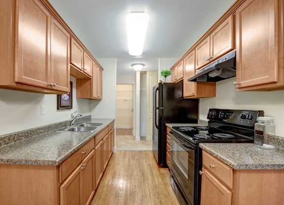 A clean galley-style kitchen here at Wyndham Crossing with warm wood cabinets, granite-look countertops, and bright overhead lighting. A double stainless sink sits along the left counter, while black appliances and plenty of storage space create a simple and functional cooking area.