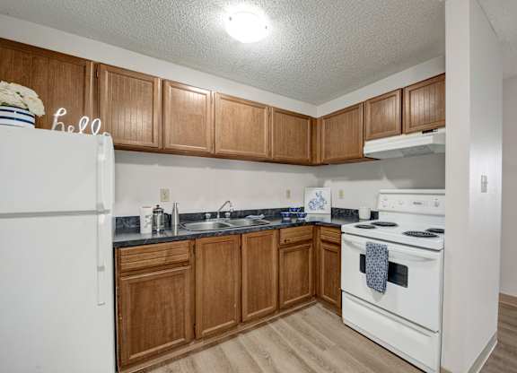 A cozy kitchen here at Wyndham Crossing with warm wood cabinets, dark countertops, and white appliances including a stove and refrigerator. A simple sink area with small countertop decor and wood-style flooring completes the clean and functional layout.
