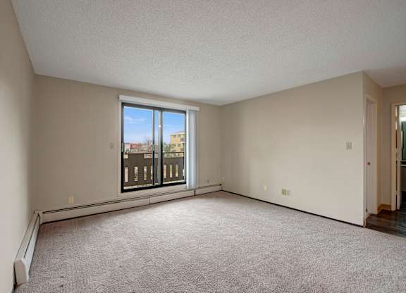 A bright living room here at Wyndham Crossing with soft carpet flooring, light neutral walls, and a wide sliding glass door that opens to a private balcony. An open layout with baseboard heating and an adjacent hallway entry creates a comfortable space for relaxing or entertaining.