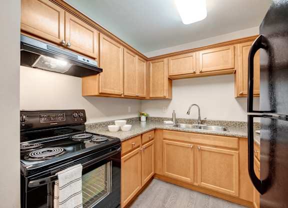 A bright kitchen here at Wyndham Crossing with warm wood cabinetry, speckled countertops, and a double-basin sink with a modern faucet. Black appliances and a smooth wood-style floor add a clean, updated look with plenty of storage and prep space.
