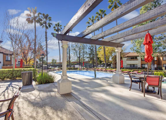 Sunny outdoor pool area here at Castlepark Apartment Homes with red umbrellas and lounge chairs. Palm trees and bushes surround the pool. A pergola casts shadows on the patio.