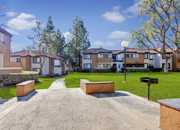 Sunlit Castleparks Apartment complex with terracotta roofs and white walls surrounds a grassy courtyard. A walkway, trees, and a barbecue grill create a serene atmosphere.