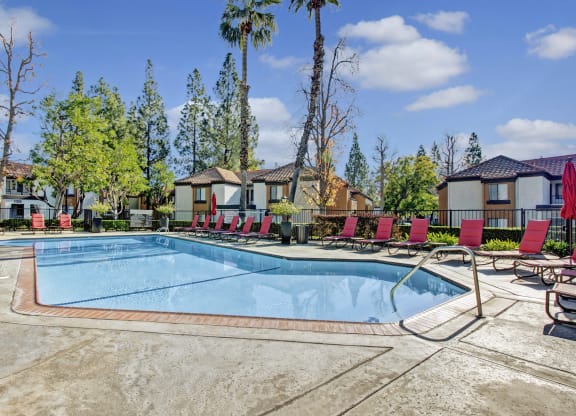 Outdoor pool area here at Castlepark Apartment Homes with clear blue water. Surrounded by several red lounge chairs and umbrellas. Trees and residential buildings are in the background.
