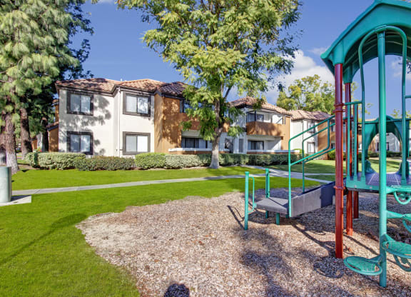 Playground here at Castlepark Apartment Homes with green jungle gym in foreground on mulch, surrounded by lush grass and trees, with beige two-story residential buildings in background.
