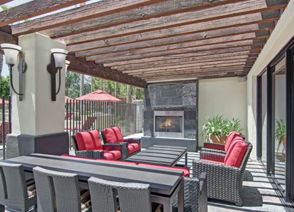 Outdoor patio here at Natura Villas Apartments with a wooden pergola, featuring red-cushioned wicker chairs, a black table, a modern stone fireplace, and potted plants. Sunny and inviting.