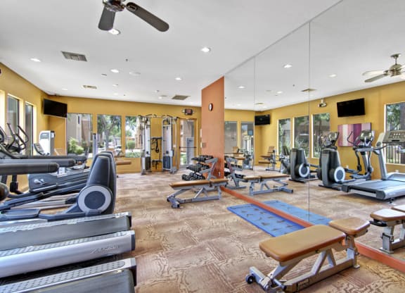 Bright gym interior here at Natura Villas Apartments with treadmills, stationary bikes, and weight benches. Mirrors reflect equipment. Large windows offer a view of greenery, creating an inviting and spacious atmosphere.