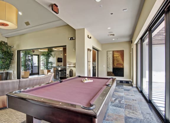 Modern recreation room here at Natura Villas Apartments with a burgundy pool table, abstract wall art, potted plants, large windows, and cozy seating. Inviting and relaxed atmosphere.