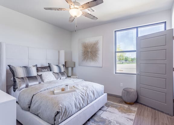Bright bedroom here at 49 West Apartments with a large bed featuring gray and silver pillows and comforter, modern art on the wall, a ceiling fan, and a sunlit window. Cozy and serene atmosphere.