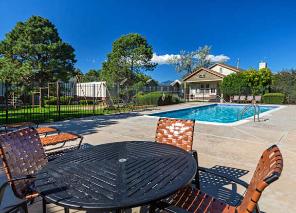 A sunny outdoor pool area here at Austin Park showcasing a sparkling rectangular swimming pool with metal handrails, a wide concrete deck, and woven patio chairs surrounding a round table in the foreground, with lounge seating, manicured greenery, mature trees, and the community clubhouse under a clear blue sky.