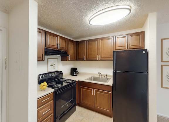 A bright, efficient kitchen here at Austin Park featuring rich wood cabinetry, black appliances including a refrigerator and electric range, light countertops, a stainless steel sink, and neutral flooring, with overhead lighting and a compact layout that offers practical storage and a clean, functional space for everyday cooking.