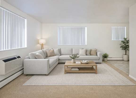 A bright modern living room here at Bristol Square featuring a spacious light gray sectional sofa with neutral throw pillows, a wooden coffee table on a soft area rug, vertical blinds allowing natural light, light-toned walls, and clean finishes that create an open, inviting atmosphere for everyday living and relaxing.