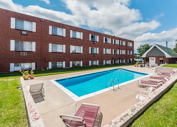 A sunny outdoor swimming pool here at Bristol Square featuring clear blue water, a rectangular pool with metal handrails, red and white lounge chairs on a concrete deck, trimmed green grass, and a brick apartment building backdrop beneath a bright sky with soft white clouds.