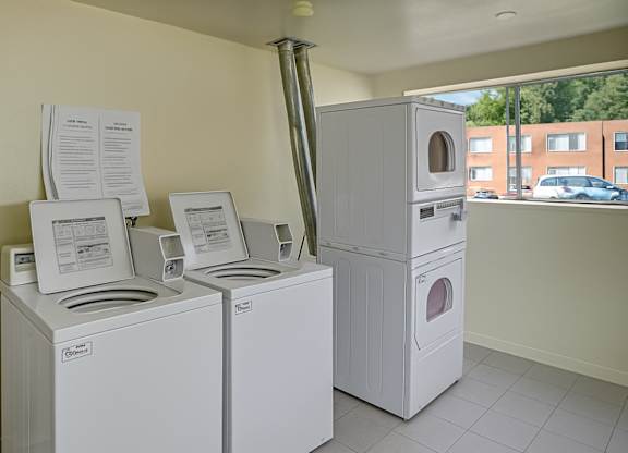 A clean shared laundry room here at Bristol Square featuring white top-load washing machines and stacked dryers, light tile flooring, neutral walls, posted laundry instructions above the machines, and a large window providing natural light and views of the apartment community outside.