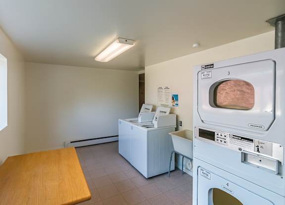 A bright shared laundry room here at Bristol Square featuring white top-load washing machines, stacked dryers, a folding table, light tile flooring, neutral walls, posted laundry instructions, and a large window that brings in natural light and overlooks the community grounds.