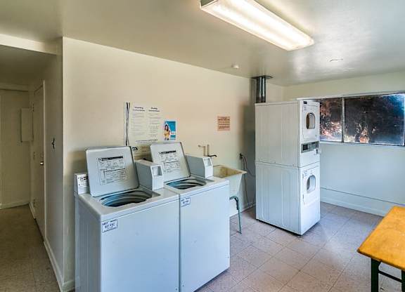 A clean, well-lit shared laundry room here at Bristol Square featuring white top-load washing machines, stacked dryers, a utility sink, a folding table, neutral walls, light tile flooring, posted laundry instructions, and overhead lighting with a window providing natural light.