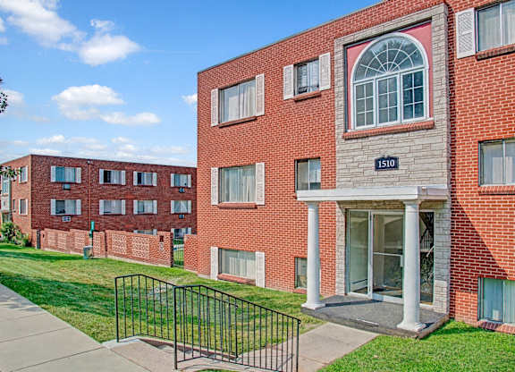 A classic brick apartment building exterior here at Bristol Square featuring a main entrance with white columns, an arched window above the doorway, multiple large windows with shutters, well-kept green lawns, a concrete walkway with railings, and additional residential buildings visible in the background under a bright blue sky.