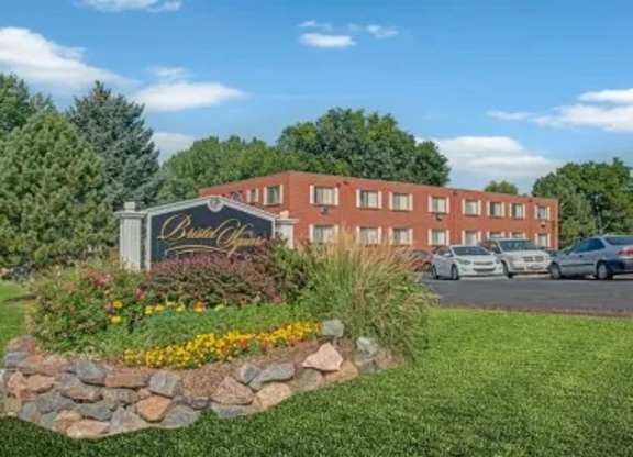 A welcoming apartment community entrance sign here at Bristol Square showcasing a landscaped monument sign surrounded by colorful flowers, decorative stone edging, green lawn, mature trees, parked vehicles, and classic brick apartment buildings beneath a bright blue sky.