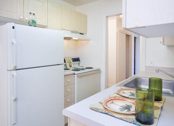 A bright and efficient kitchen here at Bristol Square featuring light wood cabinetry, white appliances including a refrigerator and electric range, smooth laminate countertops with a stainless steel sink, simple place settings with green glassware, and a compact layout that opens toward a softly lit hallway, creating a clean and functional cooking space.