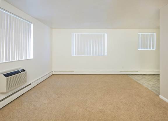 A spacious and sunlit living room here at Bristol Square showcasing soft neutral walls, plush carpet flooring, multiple wide windows with vertical blinds, a wall-mounted air conditioning unit, and an open layout that transitions into an adjacent dining area with durable flooring, creating a bright and versatile living space.