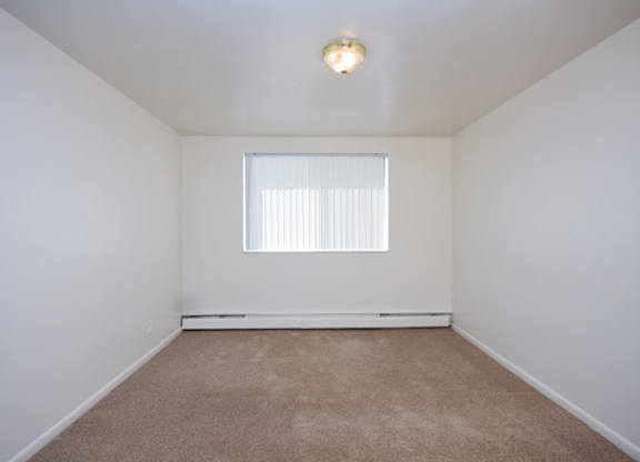 A bright and cozy bedroom here at Bristol Square featuring soft neutral walls, plush carpet flooring, a wide window with vertical blinds that allows natural light to fill the room, a simple ceiling light fixture, and a clean rectangular layout that offers flexibility for bedroom furniture and comfortable everyday living.