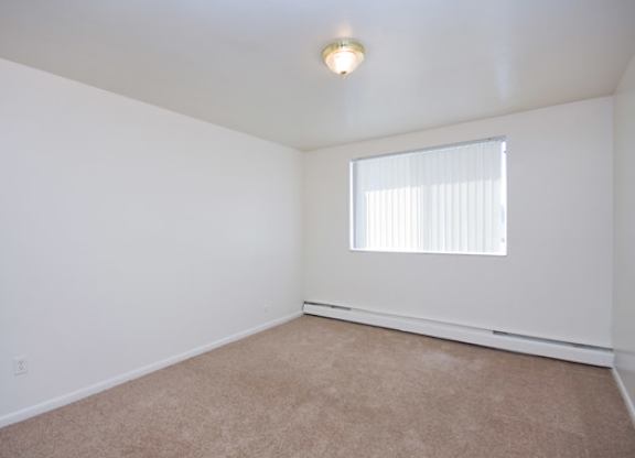 A bright and airy bedroom here at Bristol Square featuring soft white walls, plush carpet flooring, a wide window with vertical blinds for natural light, baseboard heating along the wall, and a simple ceiling light fixture, creating a clean and comfortable space suitable for a variety of bedroom furniture layouts.