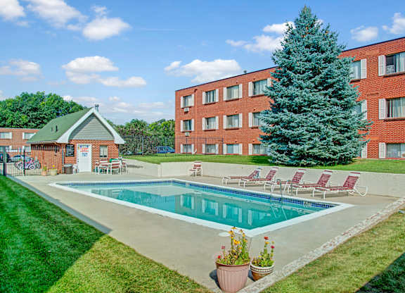 A sunny outdoor swimming pool here at Bristol Square featuring a rectangular pool surrounded by a concrete deck, red lounge chairs, green lawn landscaping, and a tall evergreen tree set against brick apartment buildings and a bright blue sky with scattered clouds.