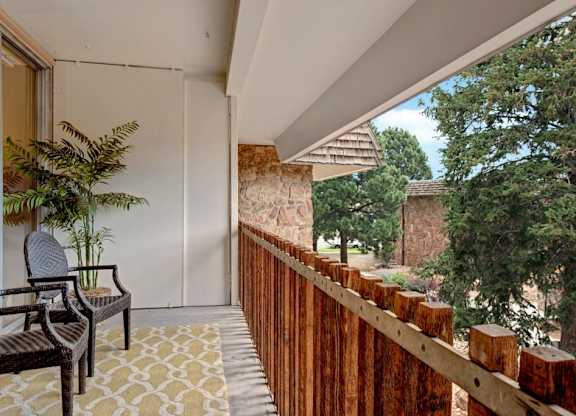 A private balcony here at Copper Stone featuring a cozy seating area with woven chairs, a patterned outdoor rug, and a potted plant, framed by a wood railing and stone accents, with peaceful views of mature trees that create a relaxing outdoor extension of the apartment.