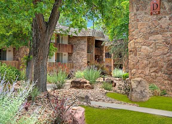 A tranquil landscaped courtyard here at Copper Stone featuring stone-accented apartment buildings with private balconies, mature shade trees, desert-inspired plants, decorative boulders, and winding concrete walkways that create a peaceful outdoor setting surrounded by lush greenery.