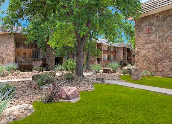 A serene landscaped courtyard here at Copper Stone showcasing stone-accented apartment buildings with private balconies, mature shade trees, desert-inspired landscaping, decorative boulders, manicured green lawn areas, and winding concrete walkways creating a peaceful residential atmosphere.