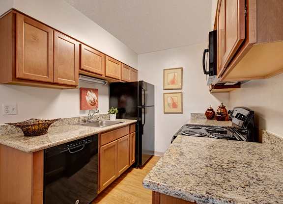 A bright, functional kitchen here at Copper Stone showcasing warm wood cabinetry, speckled countertops, black appliances, and a stainless sink, with wood-style flooring, overhead lighting, and clean finishes that create an efficient, well-organized apartment kitchen layout.