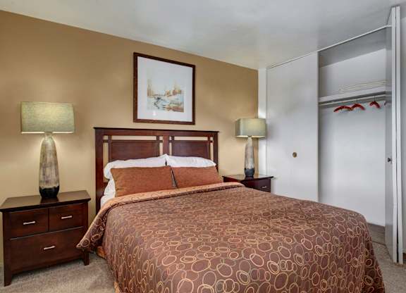 A cozy bedroom here at Copper Stone featuring a wood bed frame with patterned bedding, matching nightstands with lamps, soft carpet flooring, neutral wall tones, framed artwork above the headboard, and an open closet with shelving that adds convenient storage and a comfortable, inviting atmosphere.