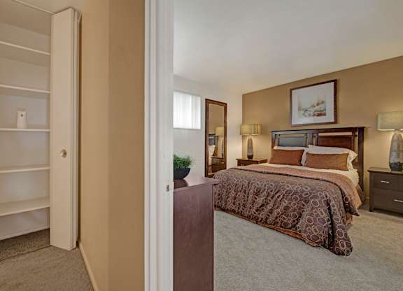 A warm bedroom here at Copper Stone showcasing a wood bed frame with patterned bedding, matching nightstands with lamps, soft carpet flooring, neutral wall tones, framed artwork above the headboard, a tall mirror, and an open shelving closet visible from the entry that adds functional storage and a comfortable, inviting layout.