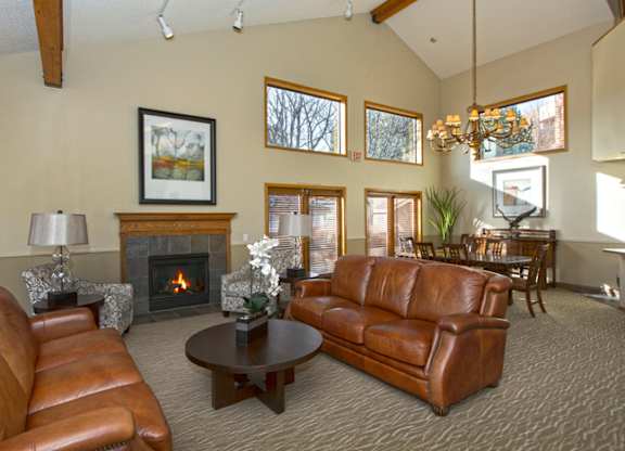 A cozy living room here at Eagle Ridge featuring leather sofas arranged around a round coffee table, a fireplace with a wood mantel, and patterned armchairs, complemented by large windows that bring in natural light, vaulted ceilings with wood beams, and an adjacent dining area with a chandelier visible in the background.