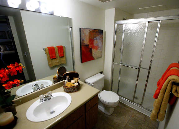 A clean bathroom here at Eagle Ridge featuring a single vanity with integrated sink and mirror, neutral countertop and cabinetry, a toilet positioned beside the vanity, a sliding glass shower enclosure with textured panels, tiled shower walls, soft overhead lighting, and neatly folded towels adding a warm accent.