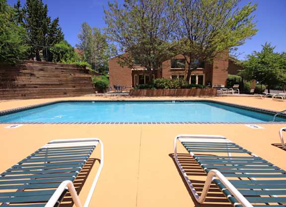 A sunny outdoor swimming pool here at Eagle Ridge showcasing a large rectangular pool with clear blue water, a wide concrete pool deck, two striped lounge chairs in the foreground, additional seating around the perimeter, landscaped trees and greenery, and a brick clubhouse building in the background beneath a clear blue sky.
