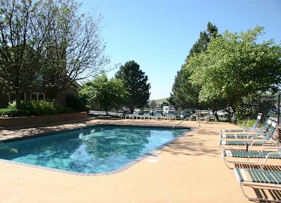 A sunny outdoor pool area here at Eagle Ridge featuring a clear rectangular swimming pool surrounded by a spacious concrete deck, neatly arranged lounge chairs, mature trees providing natural shade, and a secure fence enclosing the space, creating a relaxing and inviting community amenity on a bright day.