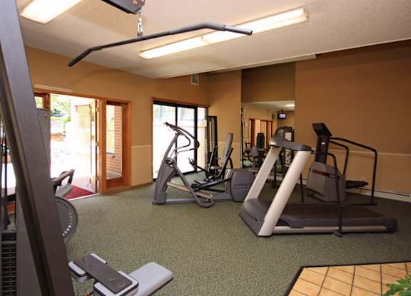 A spacious fitness center here at Eagle Ridge featuring multiple cardio machines including treadmills and an elliptical, along with strength training equipment arranged on green carpeted flooring, with overhead lighting, neutral walls, and large windows and glass doors that bring in natural light and views of the surrounding community.