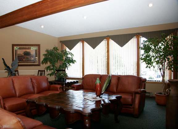 A cozy community living room here at Eagle Ridge featuring brown leather sofas arranged around a carved wooden coffee table, with large windows fitted with vertical blinds that allow natural light, exposed wood ceiling beams, neutral walls with framed artwork, and potted plants that create a warm and inviting shared gathering space.